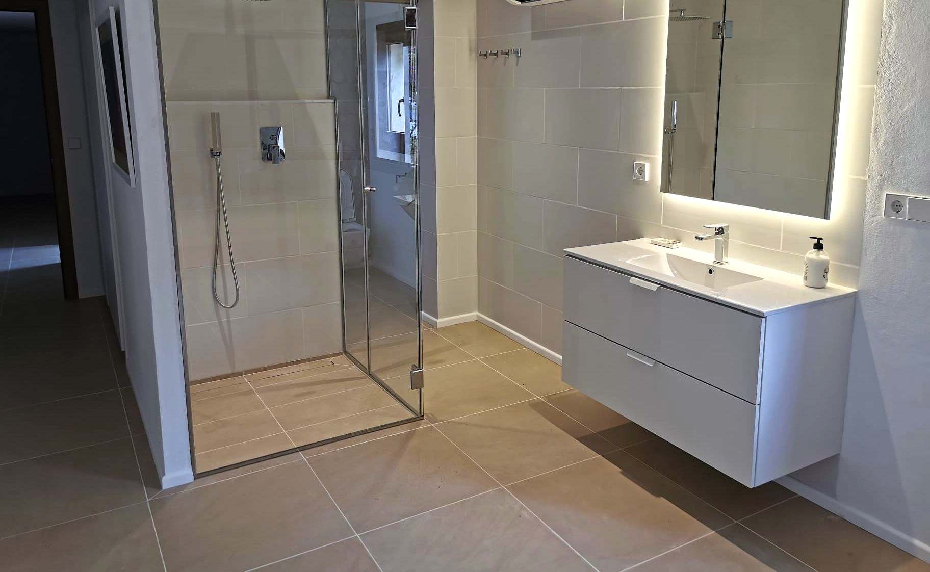 Baño blanco con mampara angular de vidrio y espejo retroiluminado sobre el lavabo